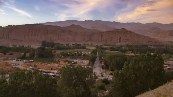 Cultivated fields and low buildings along the floor of the Bamyan valley at sunset, below bare sandstone cliffs and mountains