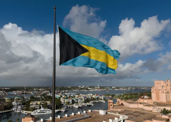 The Bahamian flag flying above Nassau harbour, with moored boats and pastel waterfront buildings below