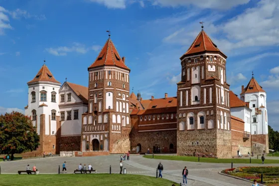 The red brick towers and white gables of Mir Castle under a blue sky, with people on the lawn in front