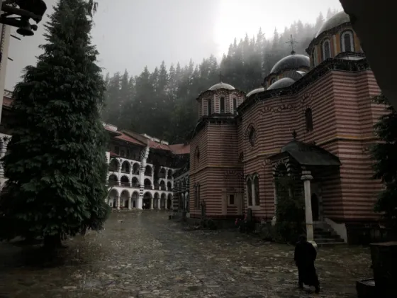 The striped arcades and domes of Rila Monastery under mist, with dark pines rising above the courtyard