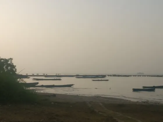 Small boats drawn up on a hazy shore of the Gambia River, with Kunta Kinteh Island low on the horizon