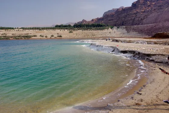 A curving pale beach along the still water of the Dead Sea, with dry cliffs rising behind