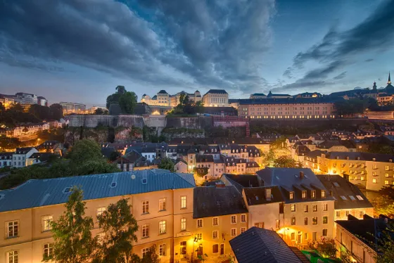 Lit rooftops of the Grund quarter below the old fortifications of Luxembourg City at dusk, under a dark sky
