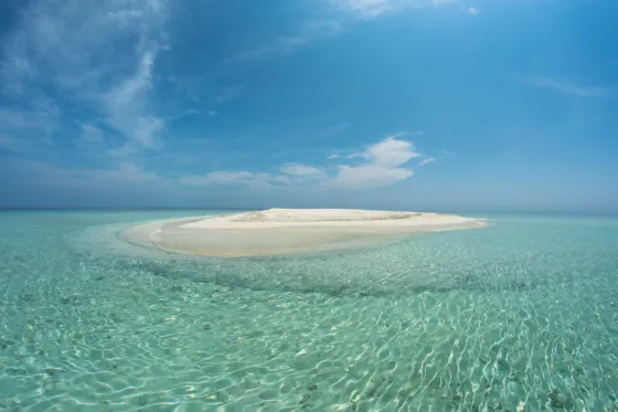 A small bare sandbank barely rising above clear turquoise shallows in the Maldives, under a wide sky
