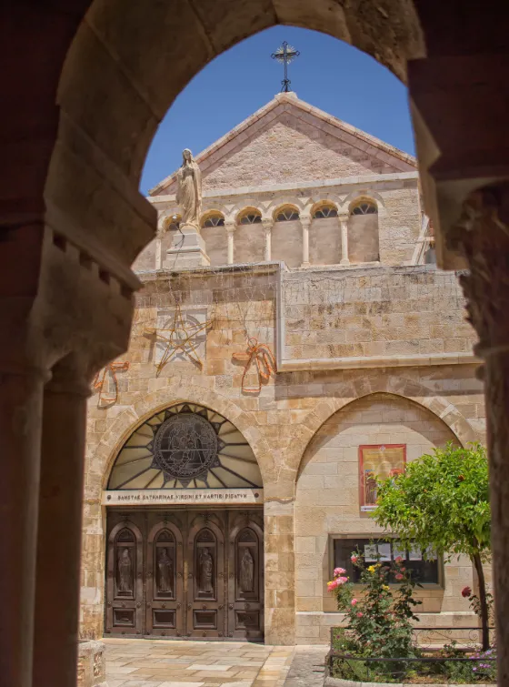 The carved stone facade of the church at Bethlehem seen through a cloister arch, with a statue above the doorway and a rose window below the gable