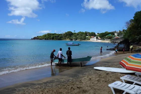 Fishermen beside a small boat on the sand at Indian Bay, with a wooded headland and houses across the water, Saint Vincent