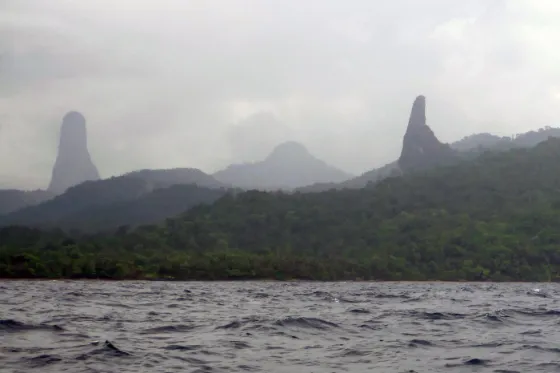 The sheer volcanic needle of Pico Cao Grande rising through haze above forest and water, Sao Tome