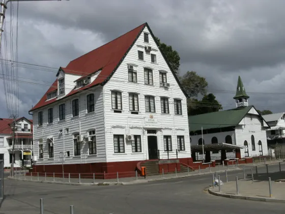 A large white timber building with a red roof on a street corner in Paramaribo, with a church beyond