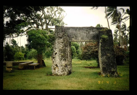 The three massive coral stone slabs of the Ha'amonga 'a Maui trilithon standing on grass beside a thatched shelter, Tonga